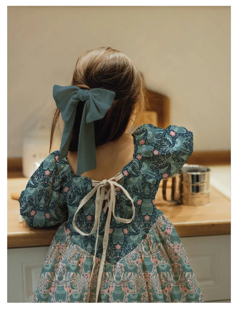 A child in a decorative dress featuring a folk animal pattern is seen from behind, with a large bow in their hair, standing by a wooden countertop.