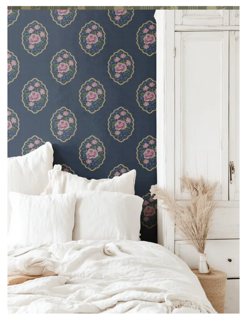 A cozy bedroom featuring a navy blue wallpaper adorned with cross-stitch roses, complemented by white bedding and decorative elements.