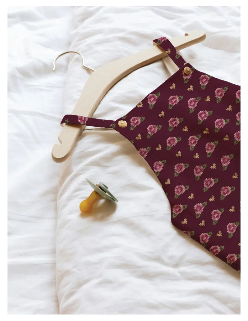 A burgundy red dress with a cross-stitch rose pattern hangs on a wooden hanger, placed on a white fabric background next to a yellow pacifier.