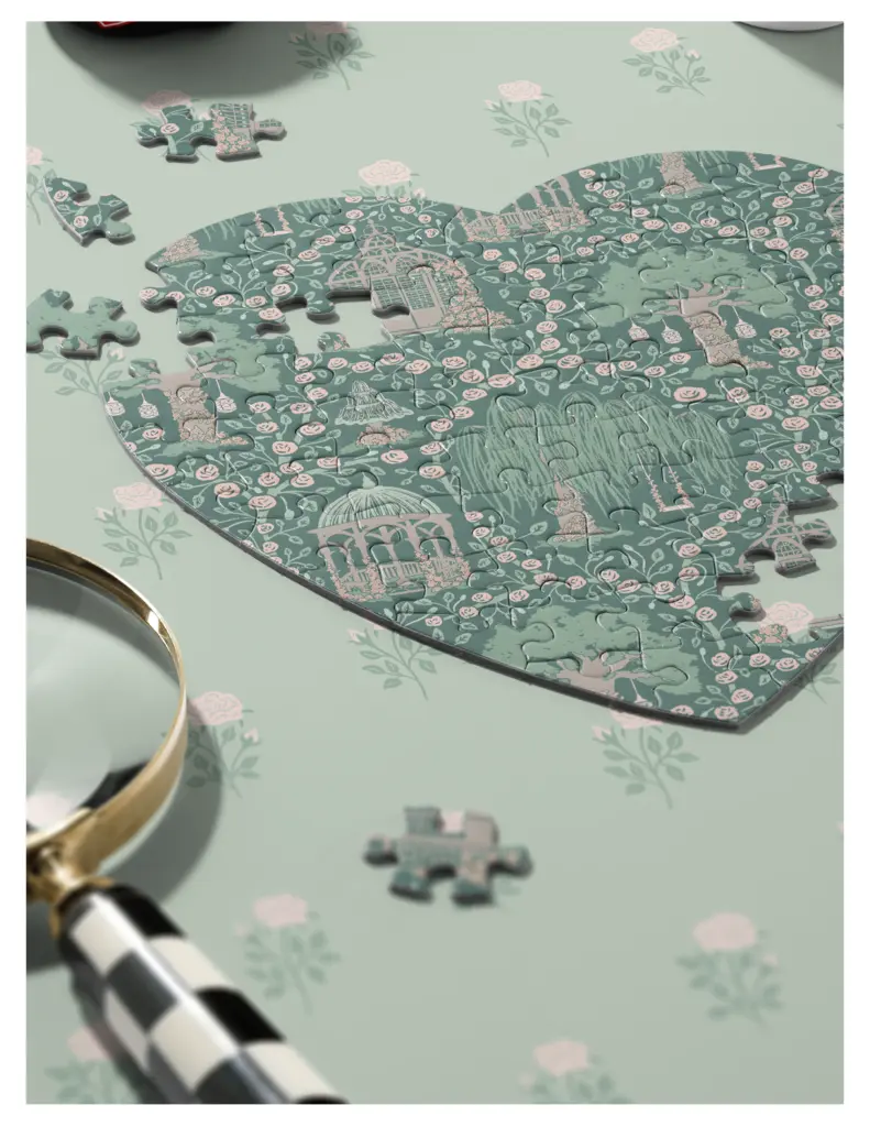 A partially completed jigsaw puzzle shaped like a heart, featuring floral and architectural designs, on a mint green table surface with roses. A magnifying glass is positioned beside the puzzle pieces.