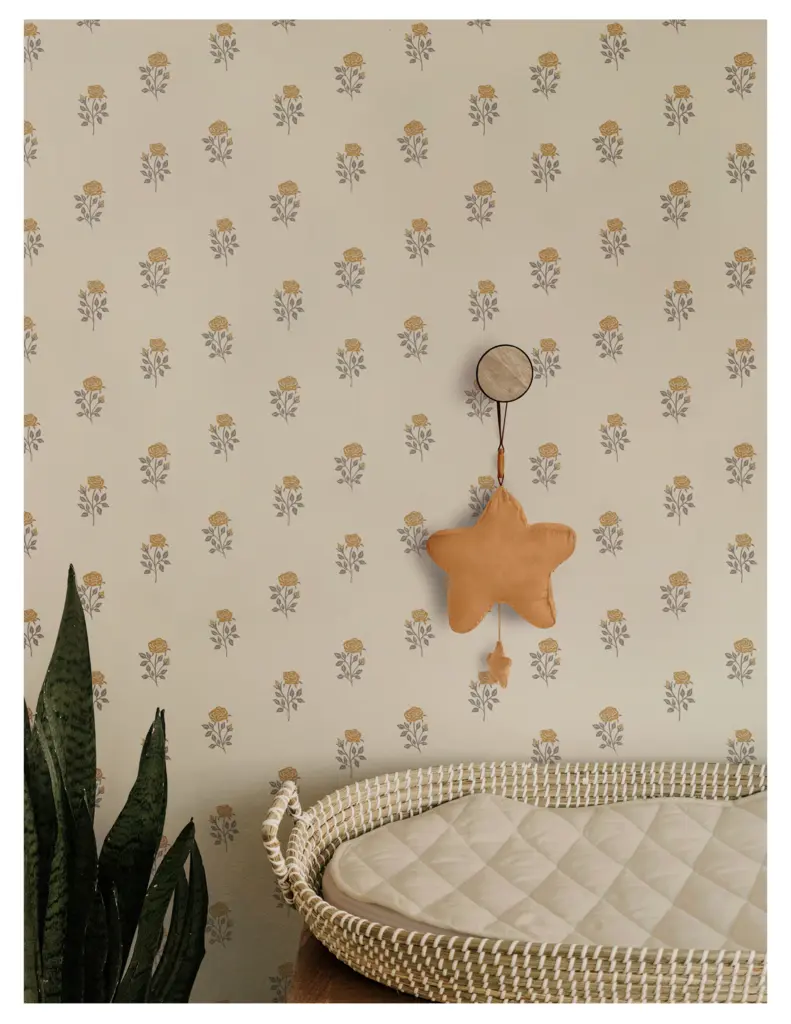 A cozy nursery scene featuring a soft beige wall with a floral pattern of yellow roses and gray leaves. There's a round mirror hanging on the wall, along with a star-shaped plush toy in a warm tan color. A woven baby bassinet with a quilted mattress is positioned in the foreground, accompanied by a green plant.