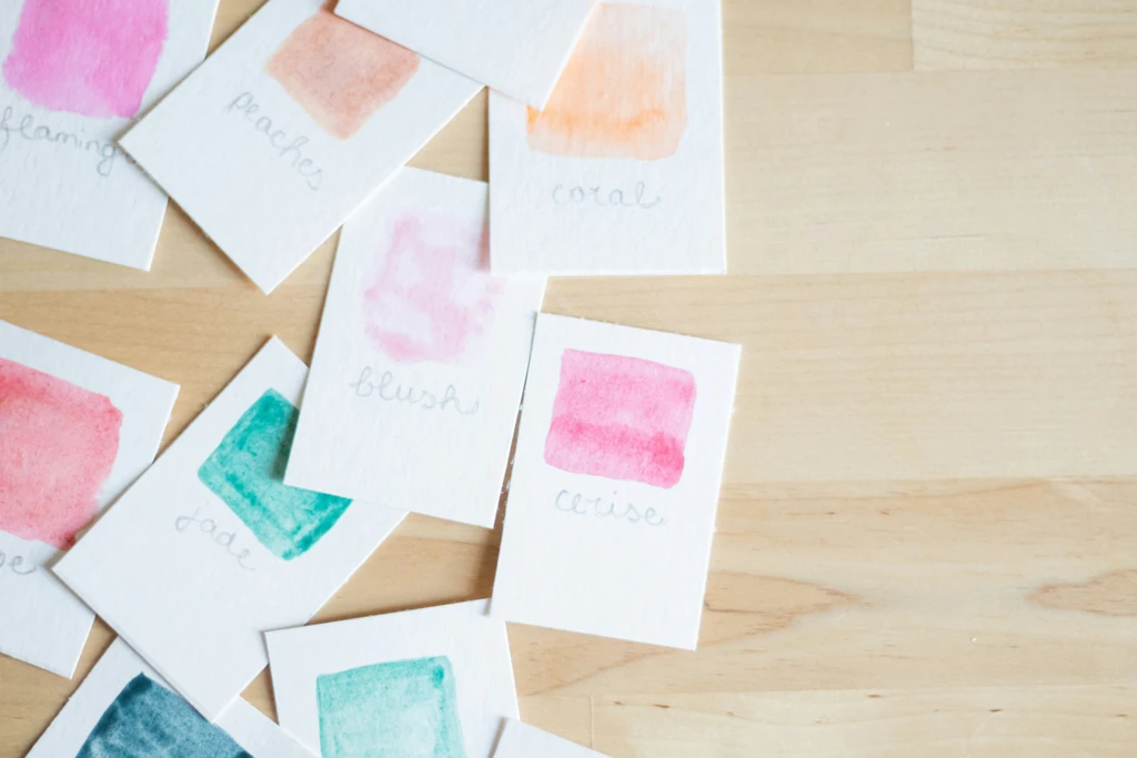 An assortment of watercolor swatches on small pieces of textured paper labeled with names of colors such as 'flamingo', 'peaches', 'coral', 'blush', 'jade', and 'cerise', arranged on a light wooden surface.