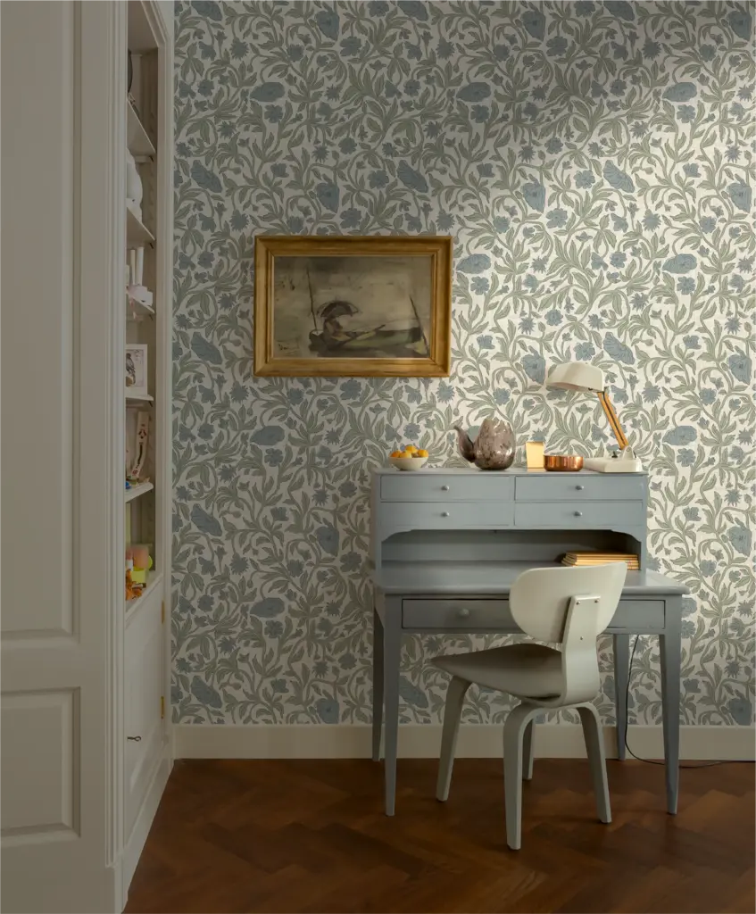 A cozy interior scene featuring a light blue wooden desk with drawers and a chair, set against a patterned floral wallpaper. A framed painting hangs above the desk, and a small lamp is positioned on it. Various decorative items, including fruit, are placed on the desk.