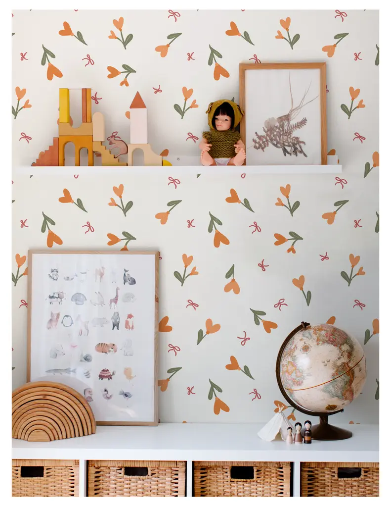 A cozy children's room decor featuring a white wall with colorful floral wallpaper. A shelf displays wooden toys, a doll dressed in a green outfit, a framed botanical print, a globe, and a wooden arch. Below, there are wicker baskets for storage.