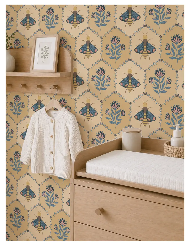 A nursery setting featuring a textured ivory cardigan hanging on a wooden hook, against a whimsical bee and floral patterned wallpaper in muted yellow tones. A light wood dresser with a changing pad sits below a small shelf holding decorative items and a plant.