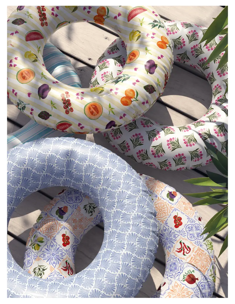 Colorful pool floats featuring various fruit patterns and floral designs, arranged on a wooden surface with green foliage in the background.