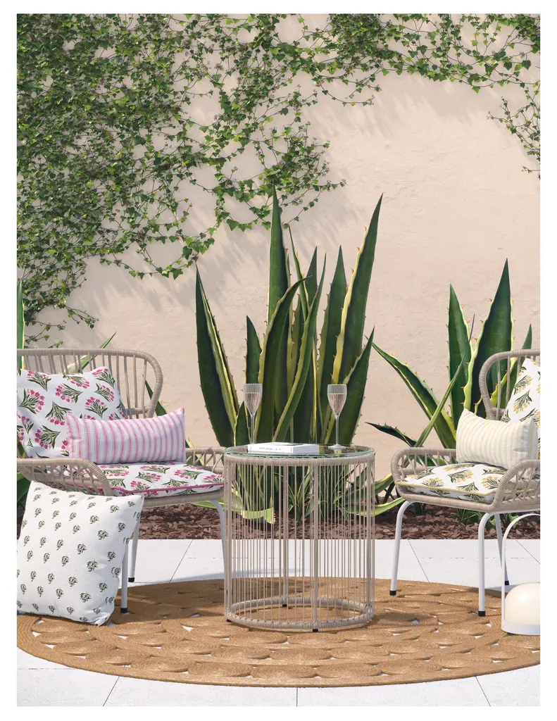 A cozy outdoor seating area featuring two stylish chairs with decorative pillows, a circular table, and a large plant backdrop. The scene includes a round jute rug and two glasses on the table.