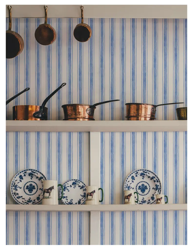 A kitchen shelf displaying several copper pots and pans hanging above, with blue and white patterned plates and mugs arranged neatly on the shelves against a striped blue wallpaper.