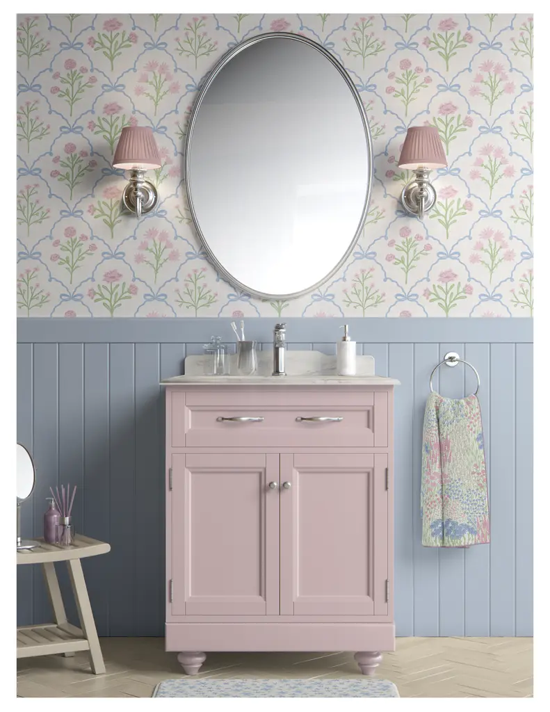 A stylish bathroom vanity in soft pink with a marble top, framed by an oval mirror and charming wall sconces. Vibrant floral wallpaper enhances the decor, while light blue paneling adds a subtle contrast.