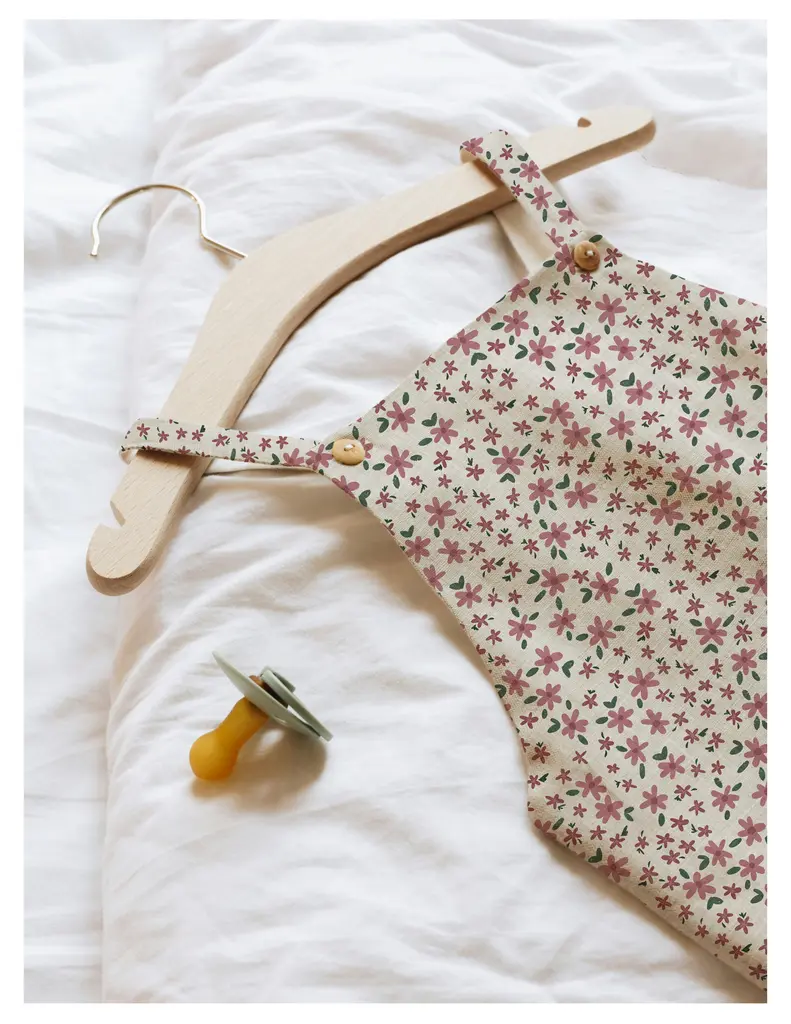A floral patterned baby outfit hangs on a wooden hanger beside a pacifier on a white textured surface.